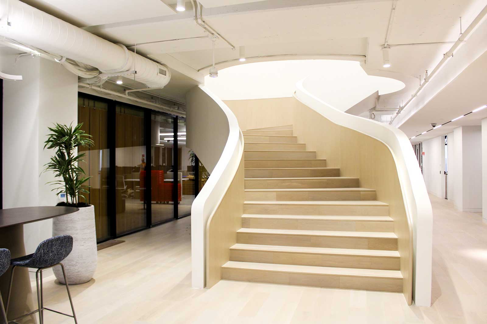 A stylish interior featuring a curved wooden staircase with white railings, surrounded by modern office furnishings and plants.