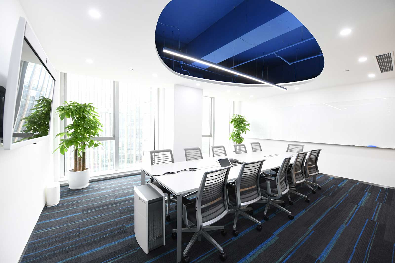 A bright, modern meeting room featuring a long table, ergonomic chairs, large windows and green plants.