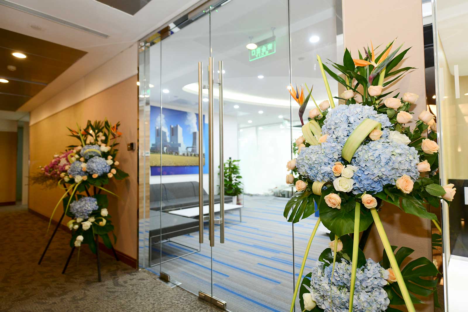 Two flower arrangements with blue hydrangeas and pale roses flank a glass door leading to a bright office space.