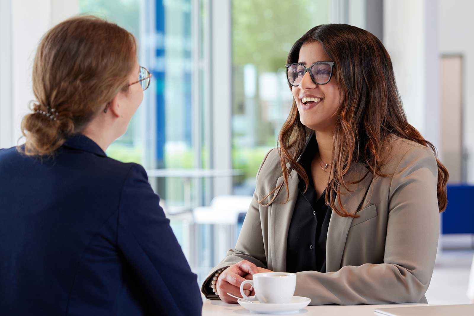 Two women engage in conversation at a bright café, one with a cup of coffee on the table.