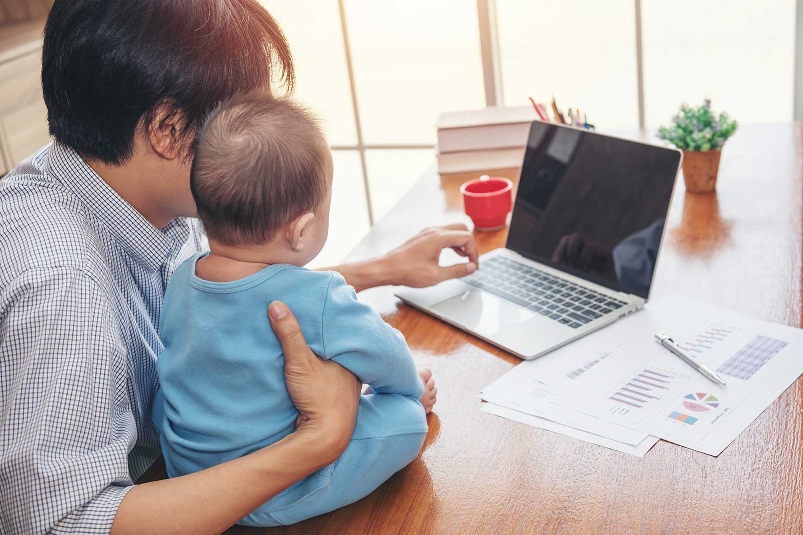 A father with black hair holds a baby in blue clothing while working on a laptop at a wooden desk with papers and a red cup.