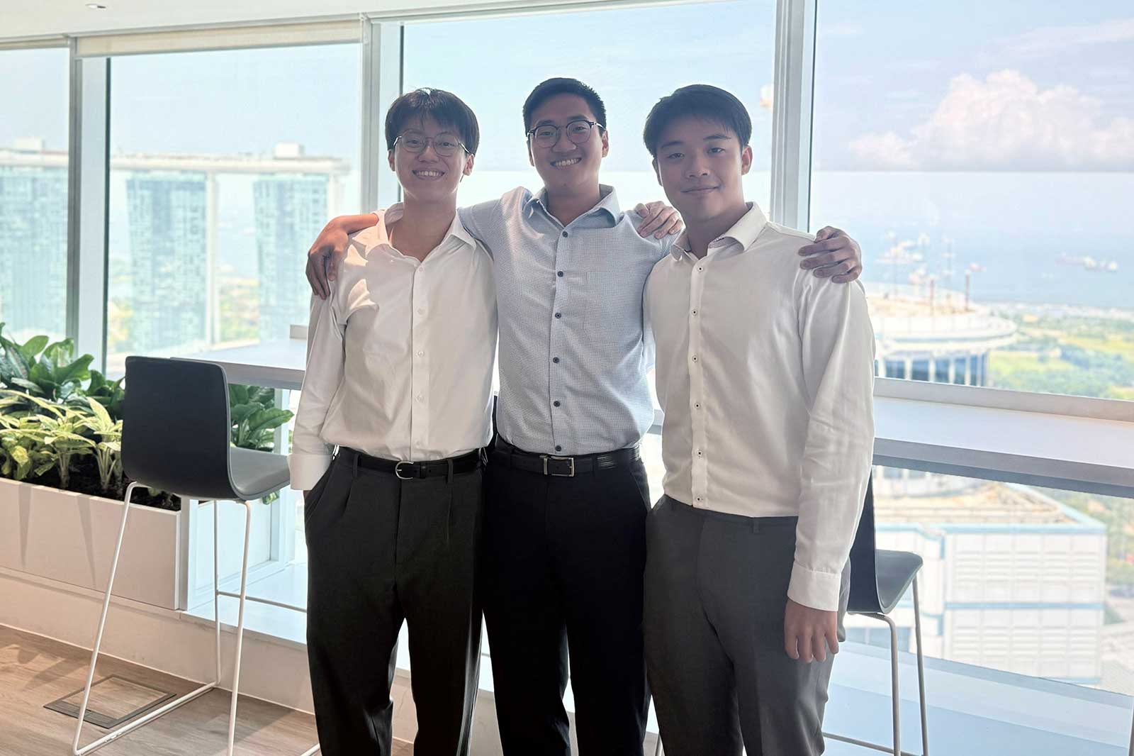 Three young men pose together in smart attire, standing by a window with a city view and greenery indoors.