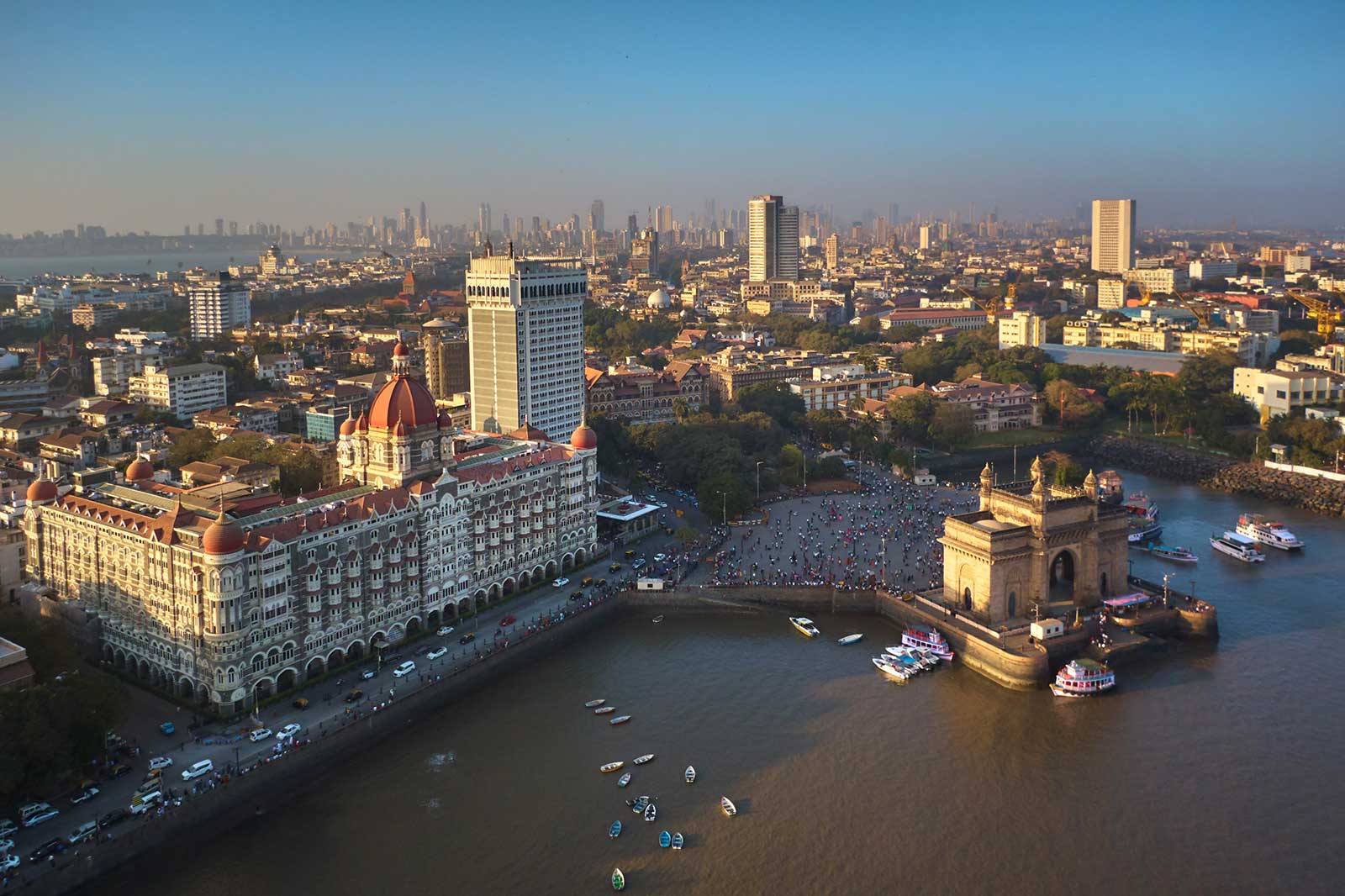 An aerial view of Mumbai featuring the Gateway of India and the Taj Mahal Palace hotel, against a skyline of modern buildings.