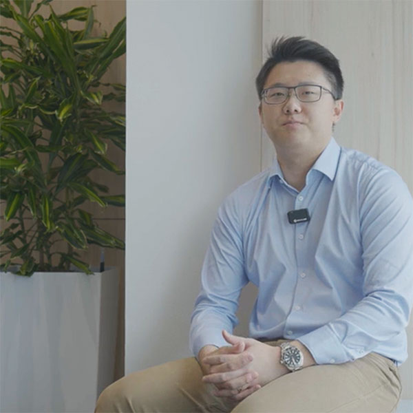 A man in a light blue shirt sits beside a green plant in a pot, with a wooden wall behind him.