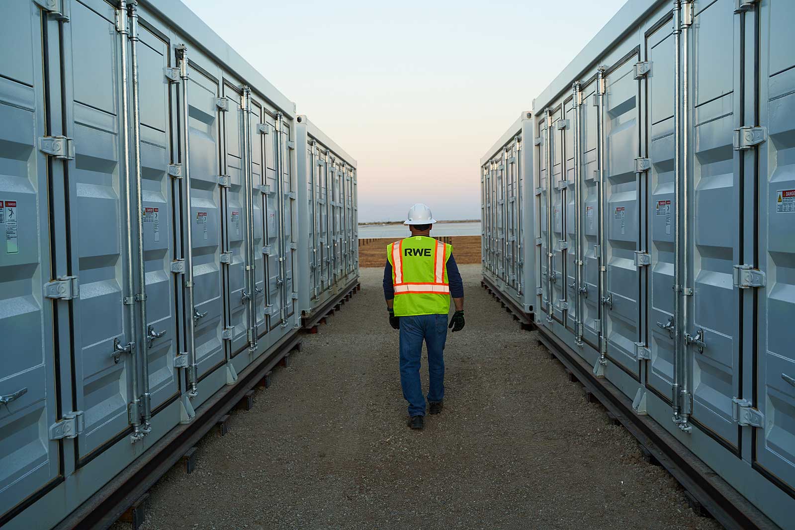 A worker in a yellow safety vest walks between two rows of grey storage containers. The evening sky is visible.