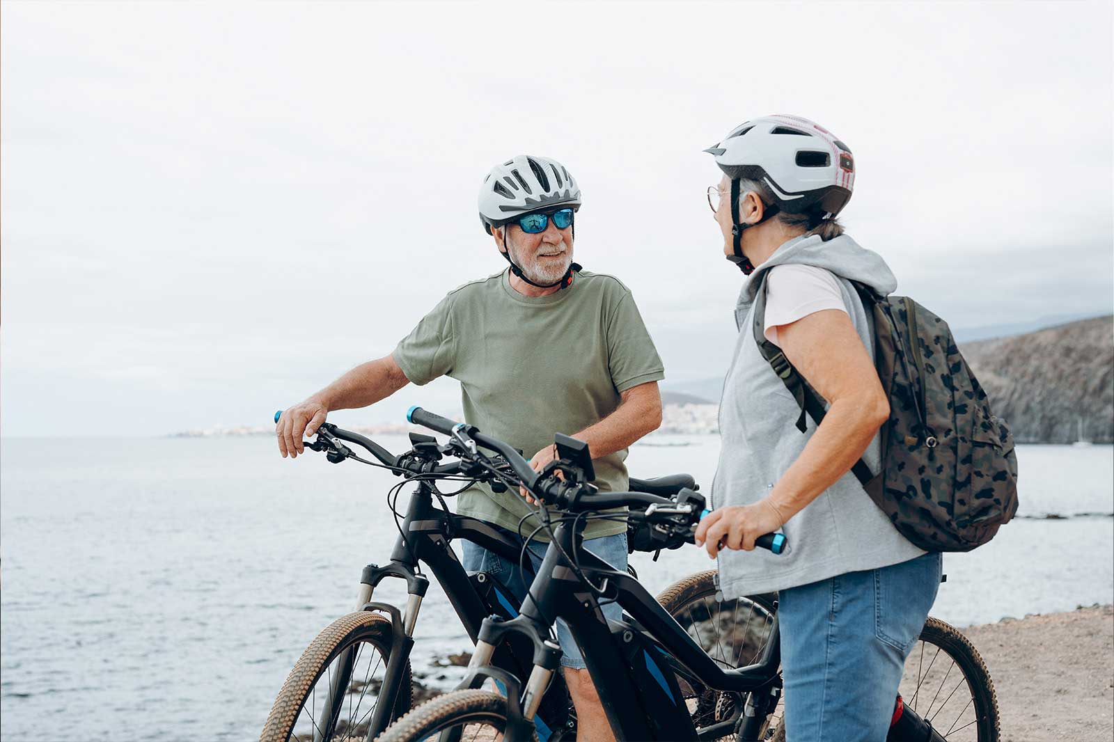 An older couple stands by a body of water, each with an e-bike, engaged in conversation. Gentle hills are visible in the background.