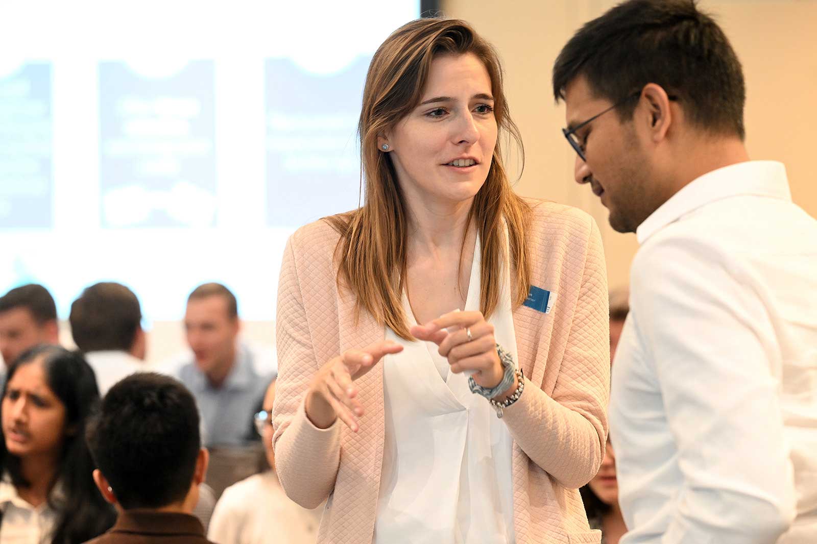 A woman is talking to a man in a conference room, while people in the background are attending an event.