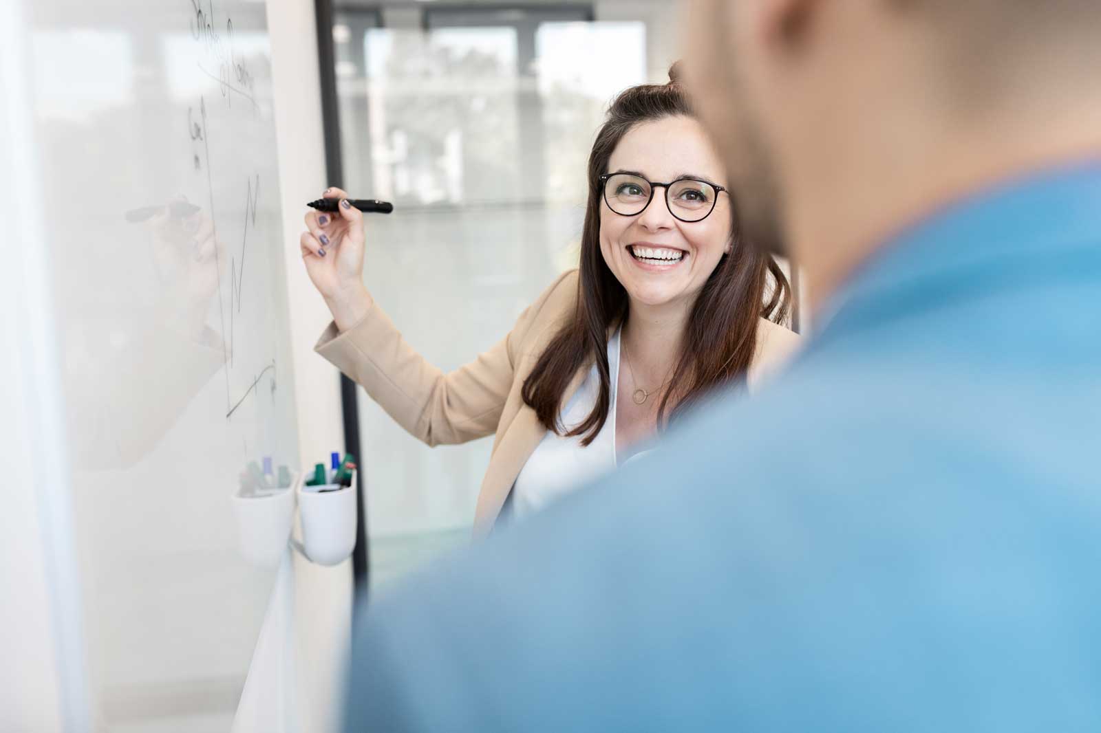 A woman presents at a whiteboard while a man listens in the foreground. She is explaining diagrams and notes.