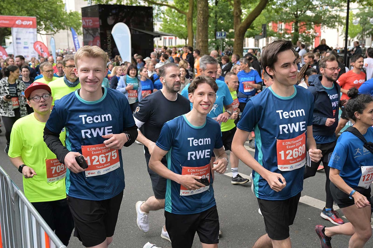 Eine Gruppe von Läufern in blauen Team RWE-Shirts nimmt an einem Stadtlauf teil. Die Umgebung ist lebhaft und grün.