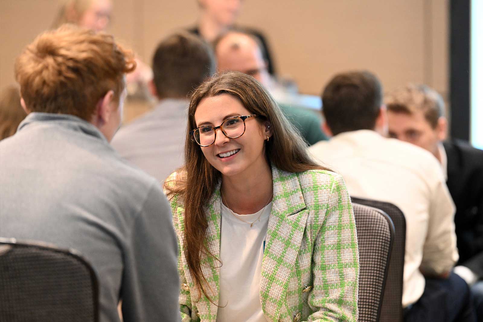 A woman with long brown hair and a checked blazer sits opposite a man in a conference room.