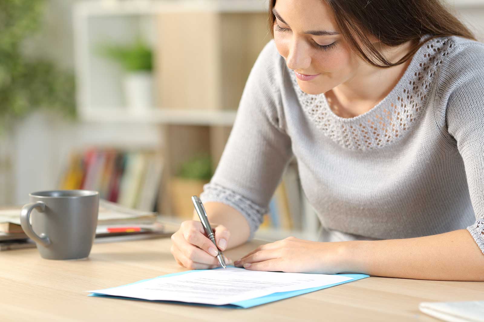 A person seated at a table is writing on a document, with a cup of coffee placed beside them.