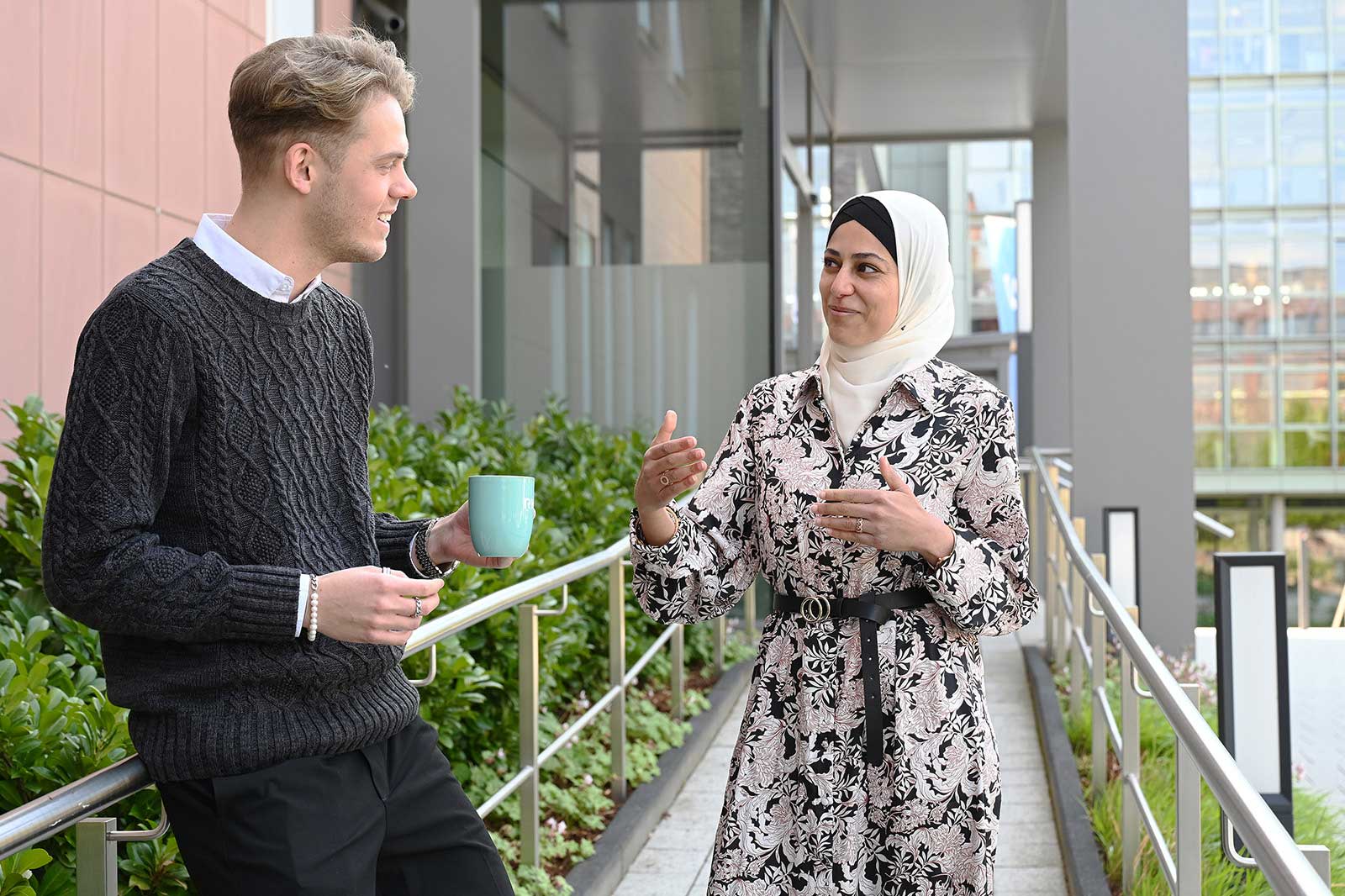 A man in a dark sweater stands holding a cup, conversing with a woman in a patterned dress.