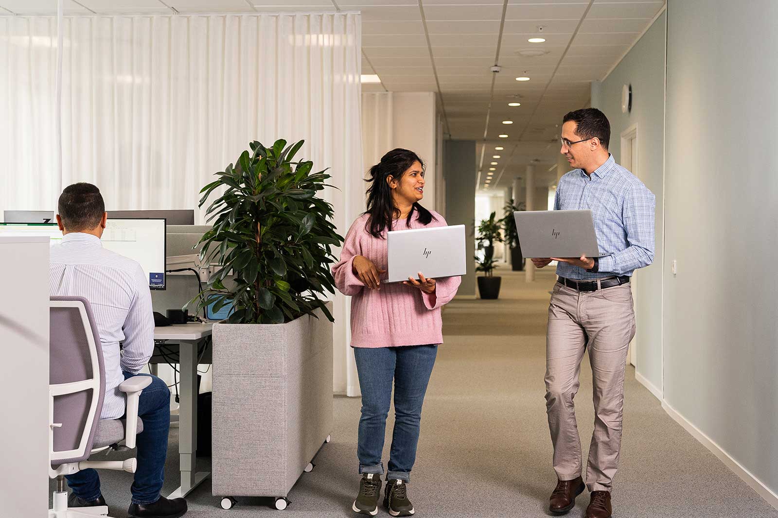 Two individuals walk in a modern office, engaging in conversation while holding laptops. Plants and workstations are visible in the background.