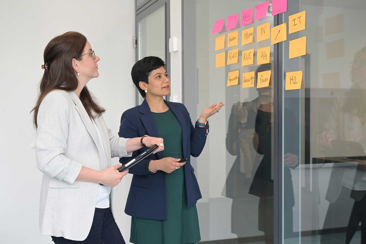 Zwei Frauen diskutieren an einer Glaswand mit bunten Notizzetteln, die auf einer Besprechung aufmerksam machen.