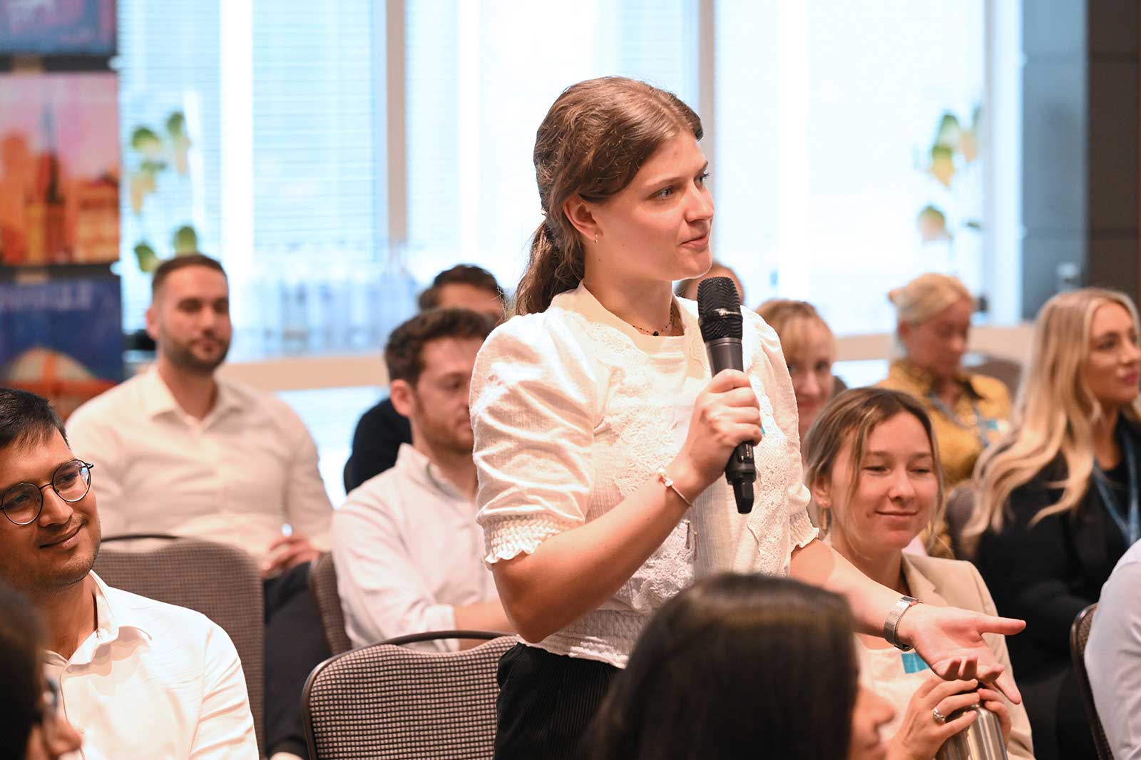 A woman asks a question at a conference, holding a microphone, with seated audience members in the background.