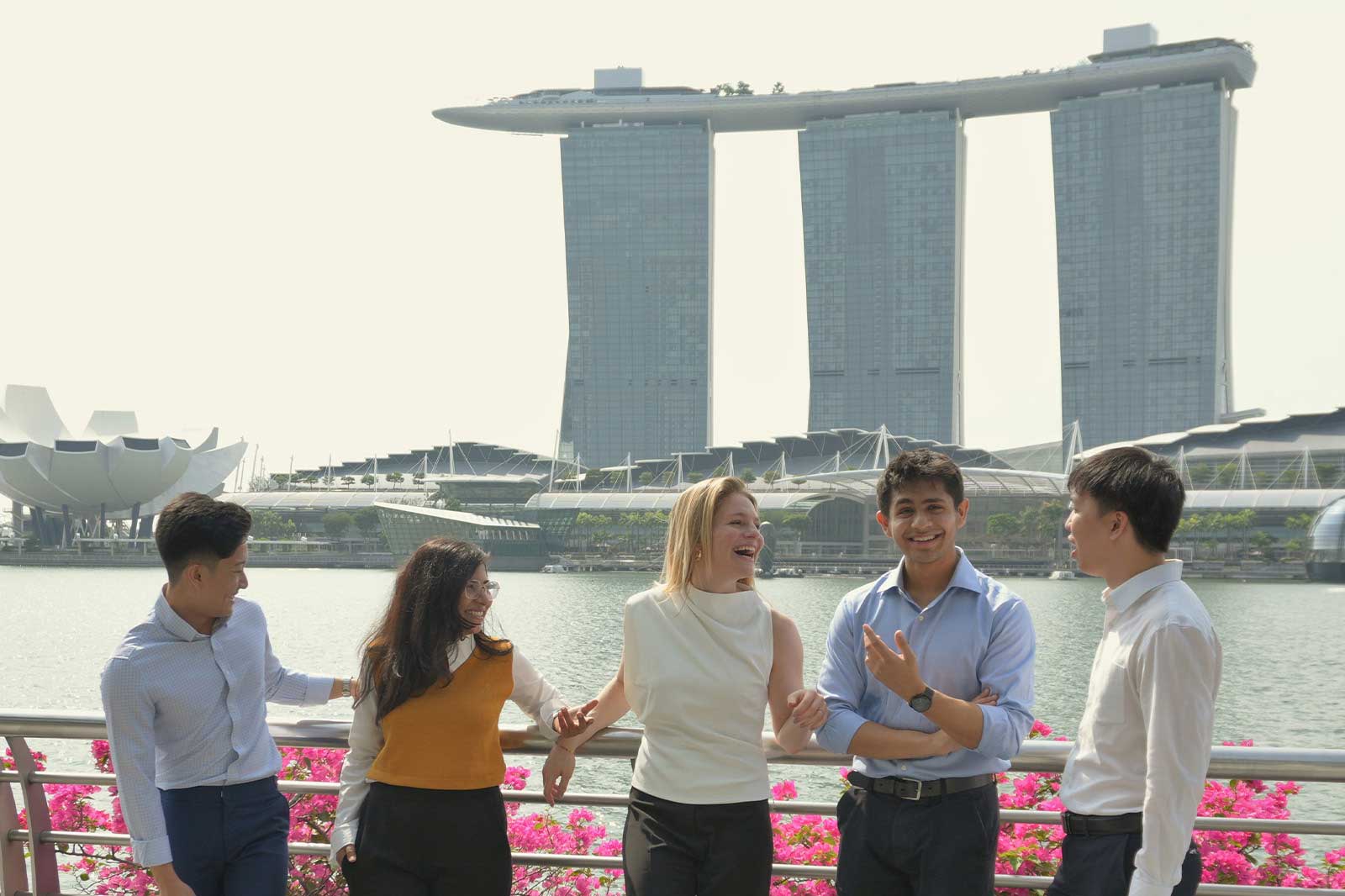 A group of five people, standing by a waterfront, with the Marina Bay Sands in the background and pink flowers in the foreground.
