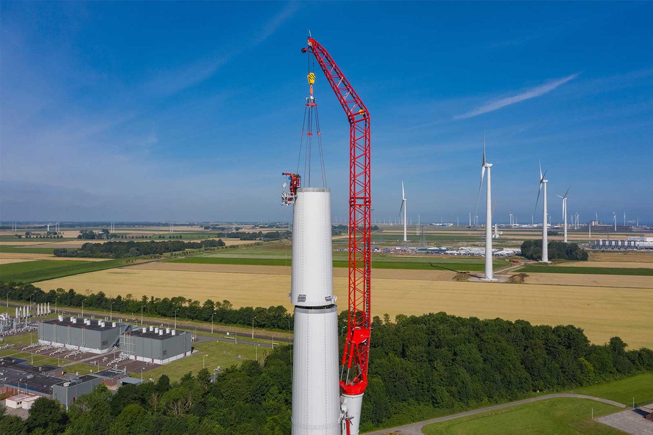 Ein Kran hebt Teile einer Windturbine in die Höhe, umgeben von Feldern und weiteren Windkraftanlagen im Hintergrund.
