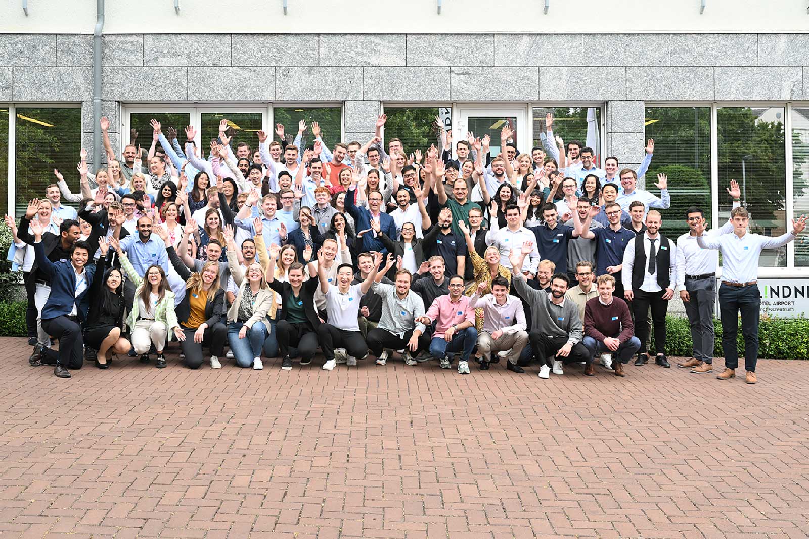 A large group of people poses in front of a building with raised hands, showcasing a cheerful atmosphere.