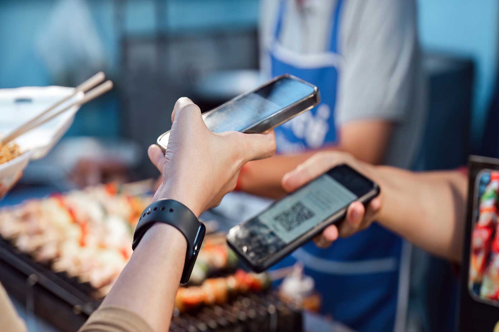 Zwei Hände tauschen Smartphones an einem Essensstand aus, eines zeigt einen QR-Code und das andere hält ein Gerät.