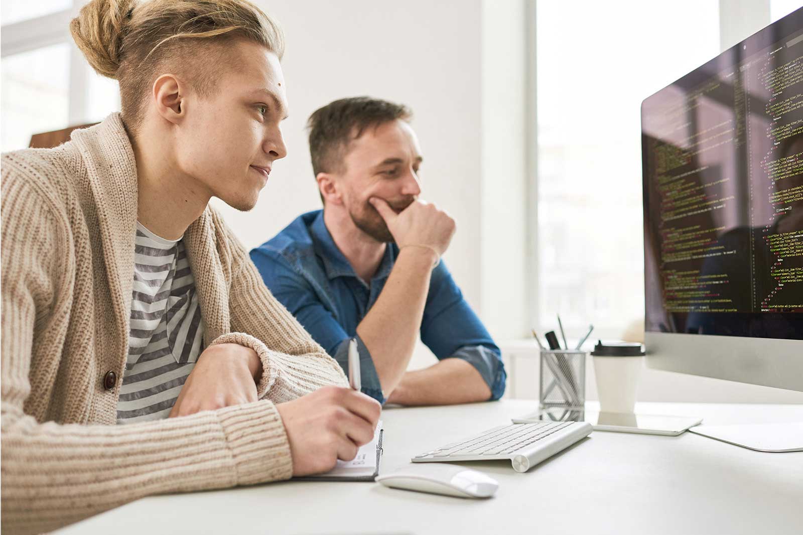 Zwei Personen diskutieren, während sie Code auf einem Computer in einer hellen Büroumgebung überprüfen.