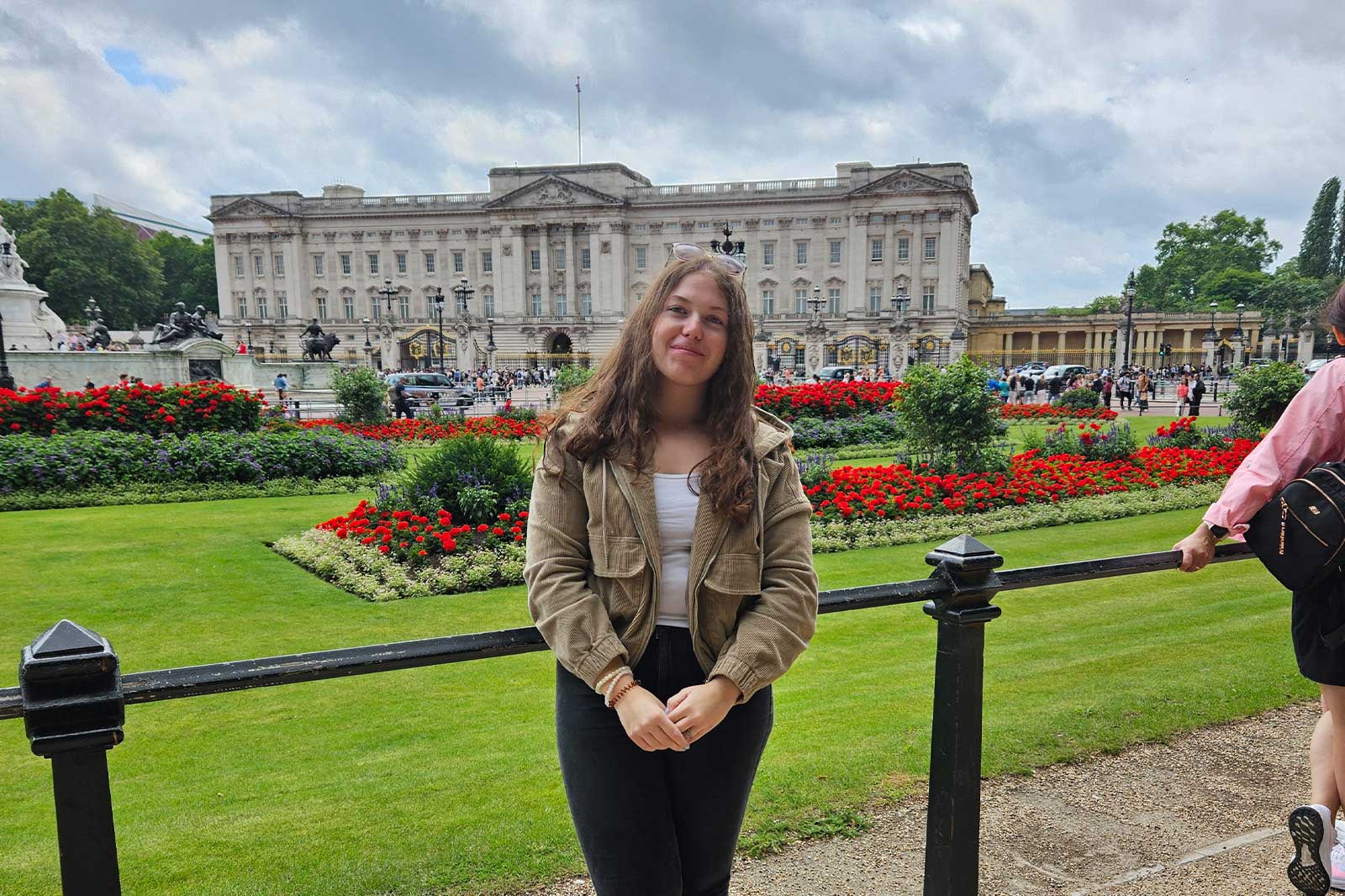 Eine junge Frau steht vor dem Buckingham Palace, umgeben von lebhaften Blumenbeeten in einem gepflegten Garten.