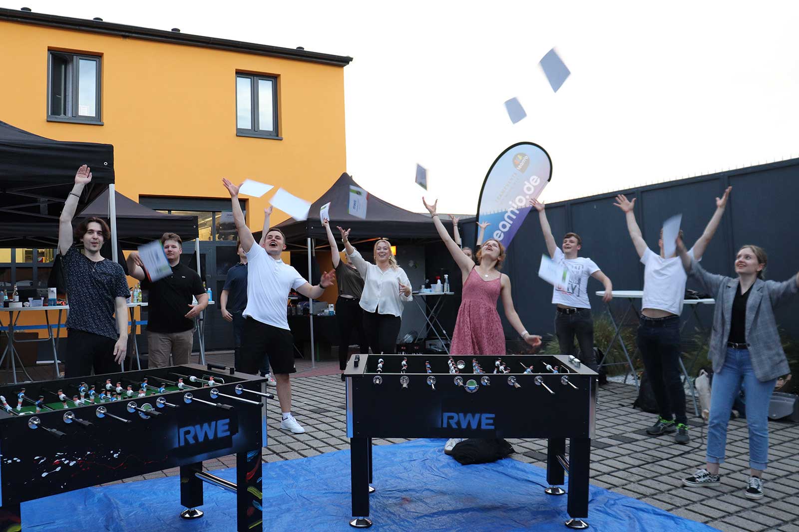 Eine Gruppe von Menschen feiert im Freien und wirft Papier in die Luft, während im Vordergrund Tischfußballtische stehen.