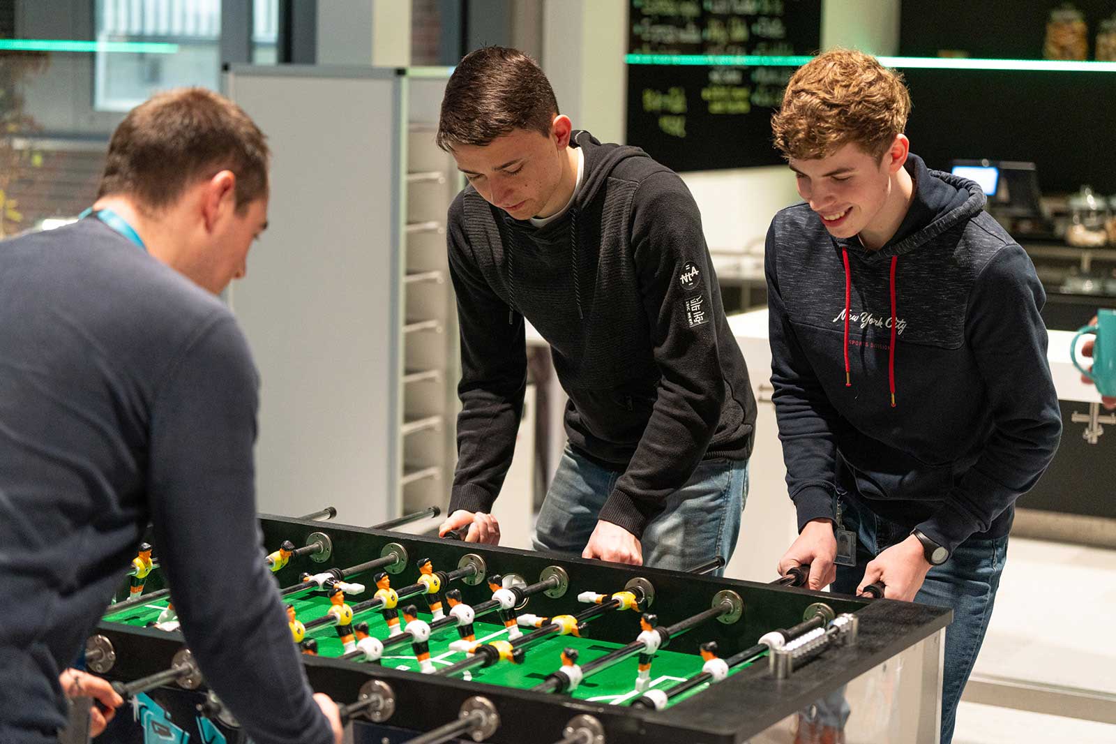 Drei junge Männer spielen gemeinsam Tischfußball in einer modernen, hellen Umgebung mit einer Kaffeemaschine im Hintergrund.