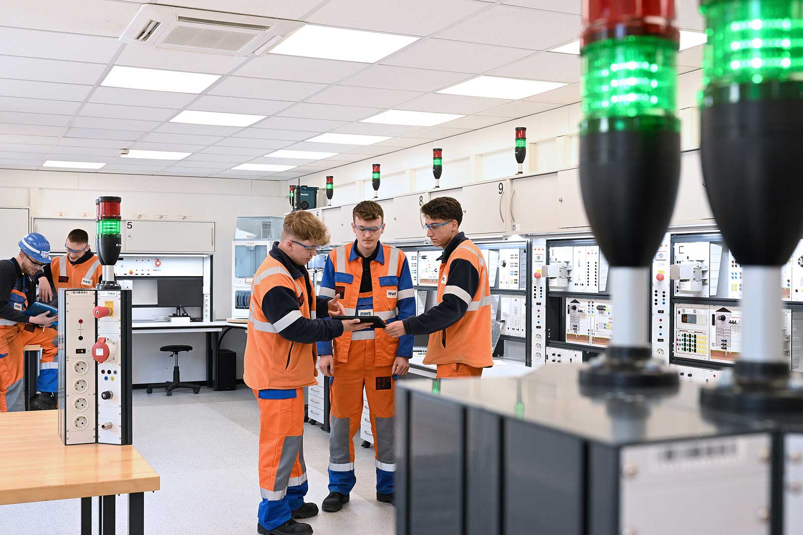 Vier Arbeiter in orangenen Sicherheitswesten unterhalten sich um ein Tablet in einem hell erleuchteten technischen Schulungsraum mit Geräten.