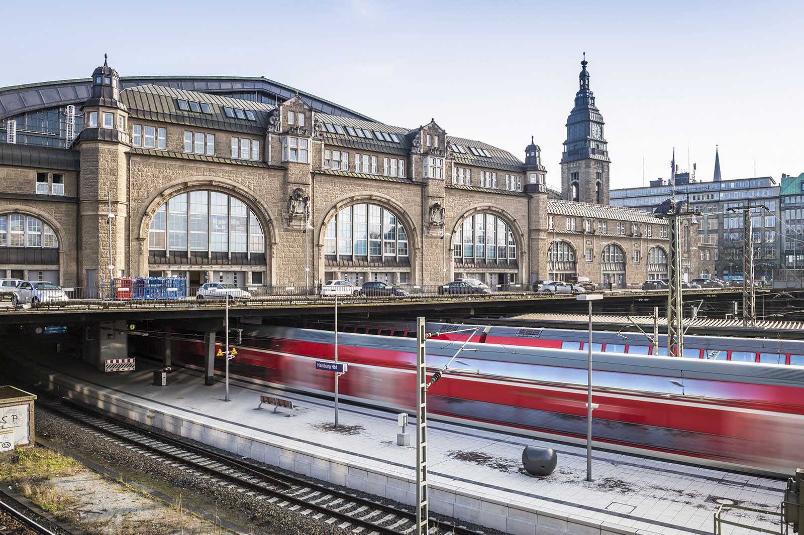 Ein roter Zug fährt an der historischen Fassade des Hamburger Hauptbahnhofs vorbei. Das Gebäude hat große Rundbogenfenster.