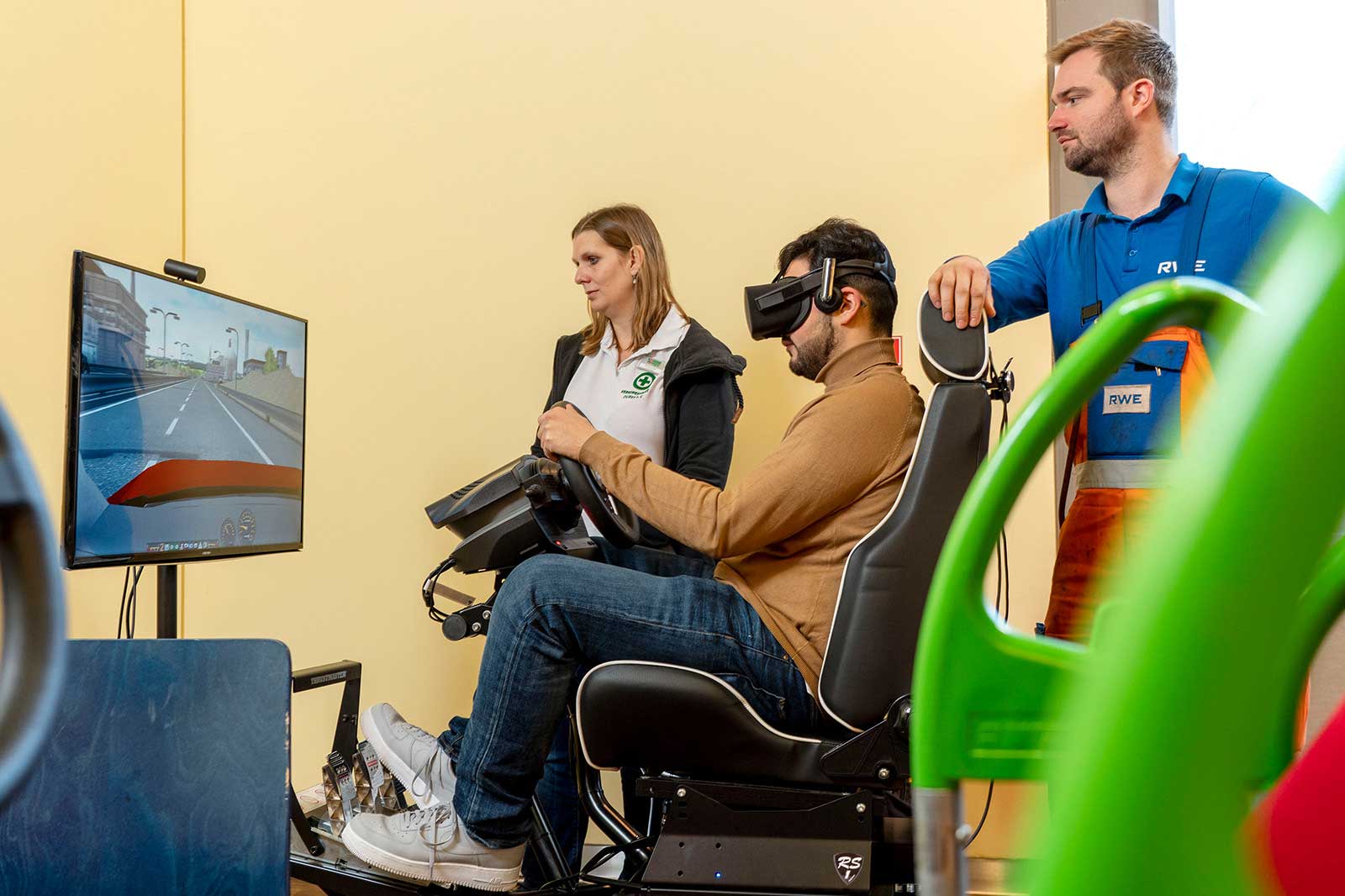Eine Person sitzt in einem Fahrsimulator und verwendet ein Virtual-Reality-Headset, während zwei andere das Erlebnis beobachten.