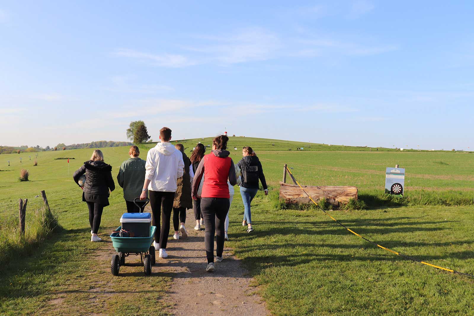Eine Gruppe von Menschen geht an einem grasbewachsenen Weg zu einem Hügel an einem sonnigen Tag, mit einem Wagen voller Vorräte.