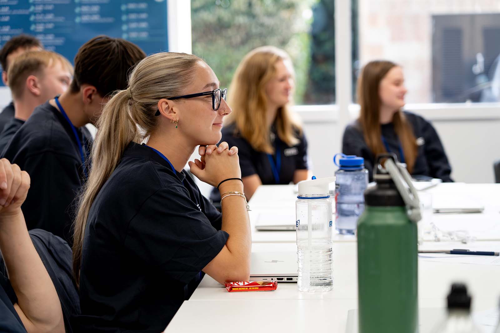 Eine blonde Frau mit Brille sitzt im Vordergrund am weißen Tisch, umgeben von Jugendlichen und Wasserflaschen, aufmerksam in einem Klassenraum.