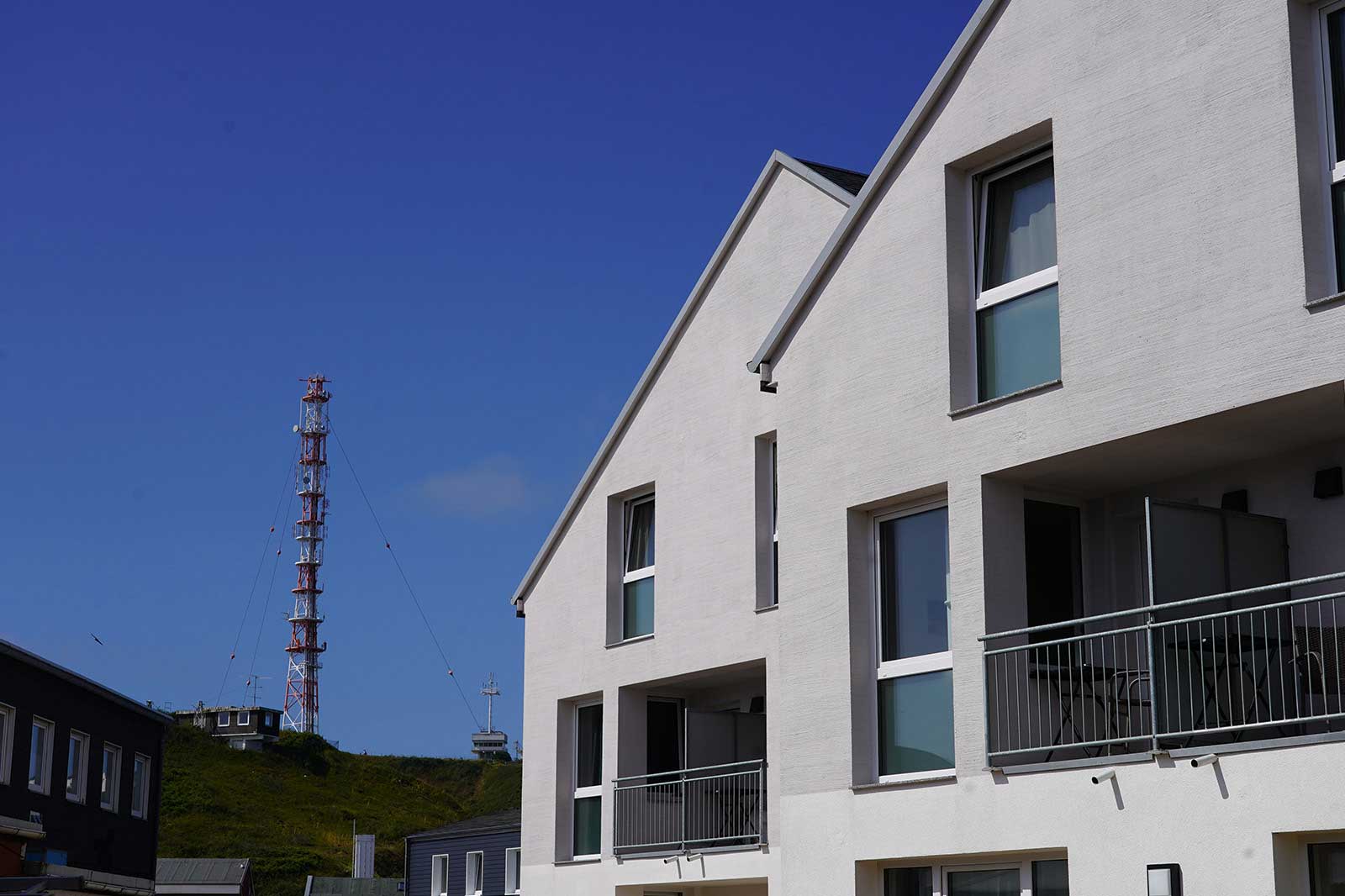 Ein modernes Wohngebäude mit einem Balkon, großen Fenstern und einem klaren blauen Himmel, im Hintergrund ein Telekommunikationsturm.