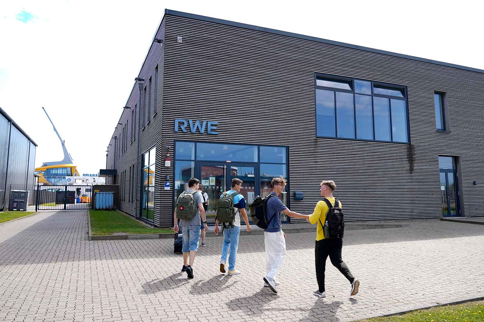 Vier junge Leute mit Rucksäcken gehen an einem sonnigen Tag auf ein modernes Gebäude mit der Aufschrift RWE zu.