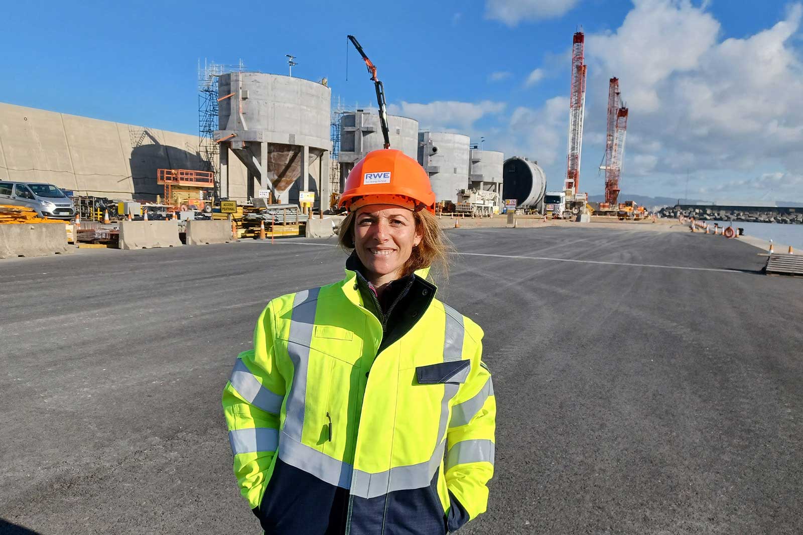 Ein Arbeiter in einer leuchtend gelben Sicherheitsjacke und orangefarbener Schutzhelm steht auf einer Baustelle mit Kränen im Hintergrund.