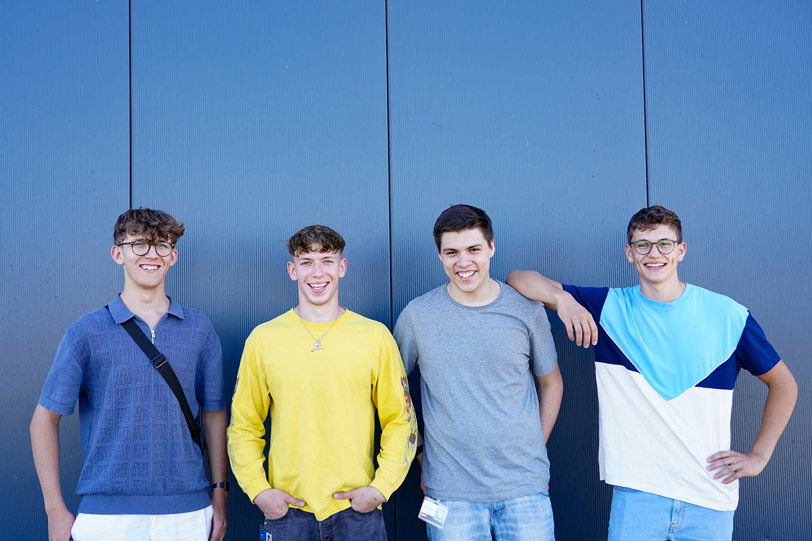 Vier junge Männer stehen vor einer blauen Wand, gekleidet in lässige Outfits, und zeigen eine freundliche und entspannte Haltung.