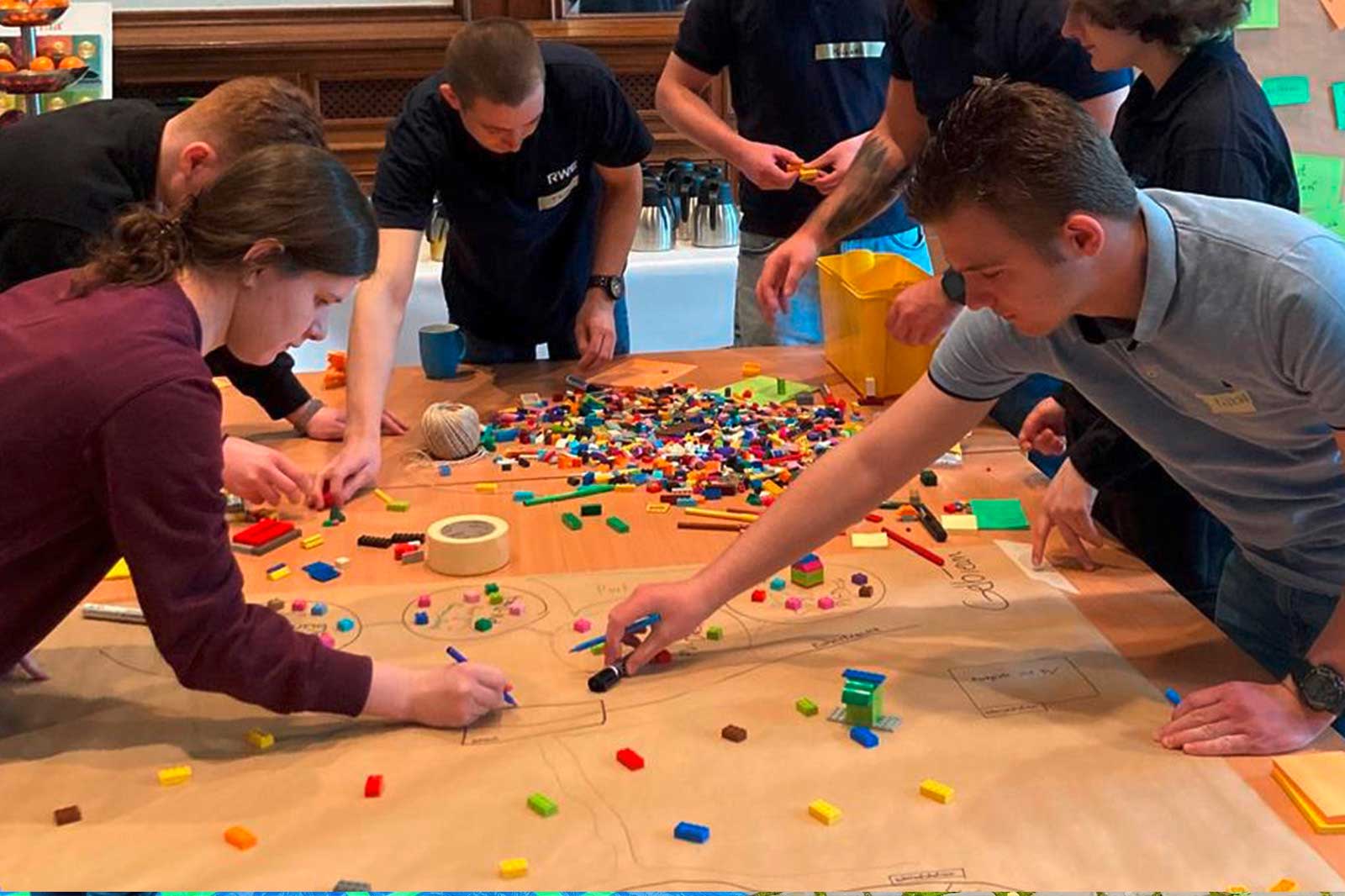 Eine Gruppe von Menschen arbeitet an einem Tisch, der mit bunten Bausteinen und Papier bedeckt ist, und beteiligt sich an einer kreativen Aktivität.