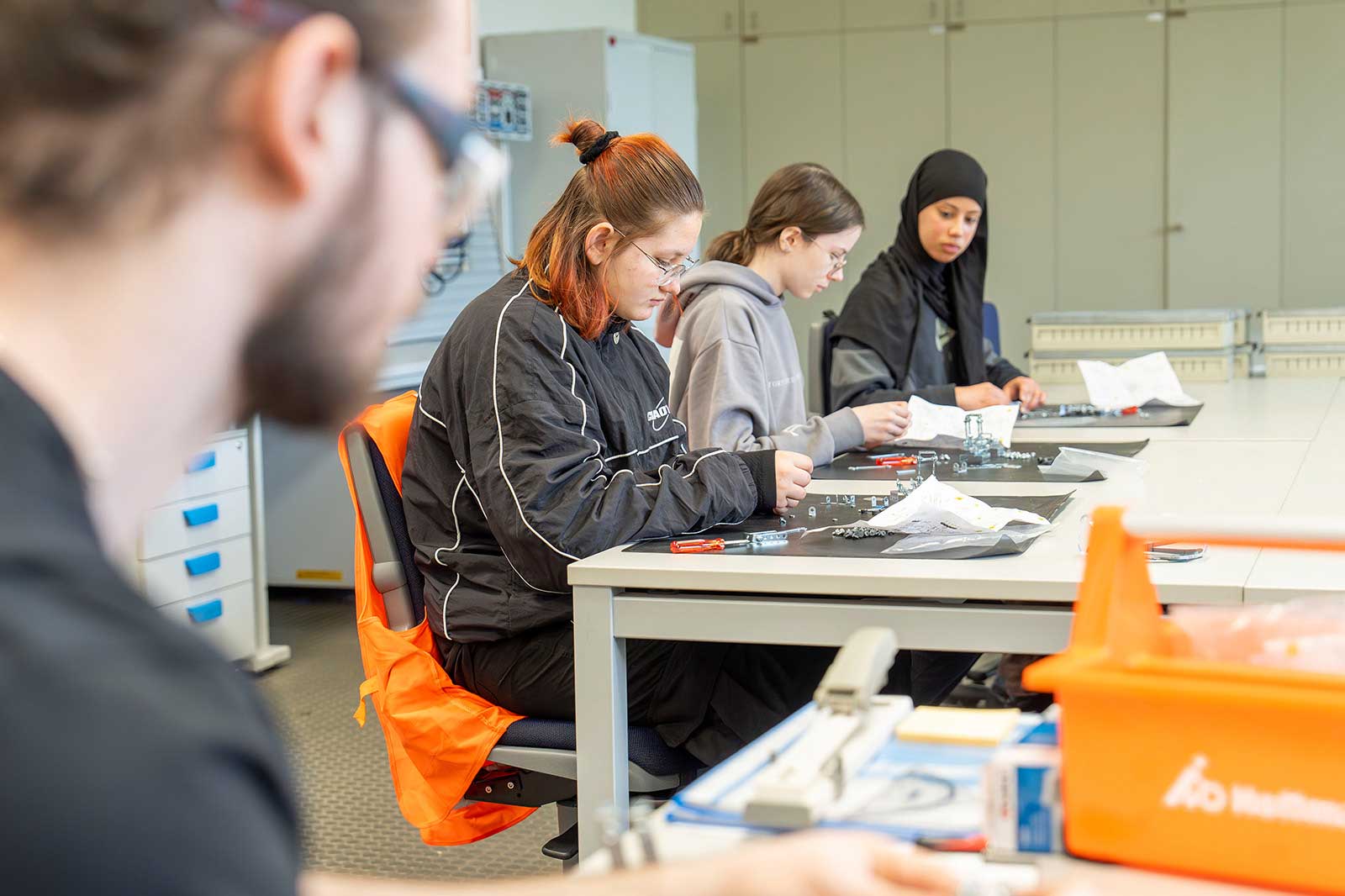 Eine Gruppe von Personen, die an einem Montageprojekt an einem Werkstatttisch arbeiten, wobei sie sich auf verschiedene Werkzeuge und Materialien konzentrieren.