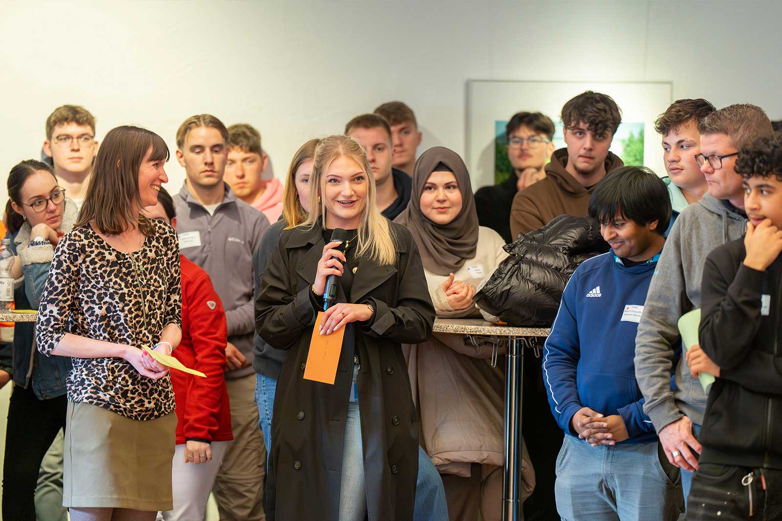 Eine Gruppe von Menschen versammelt sich um einen Tisch, während eine Frau in ein Mikrofon spricht und Informationen bei einer Veranstaltung teilt.