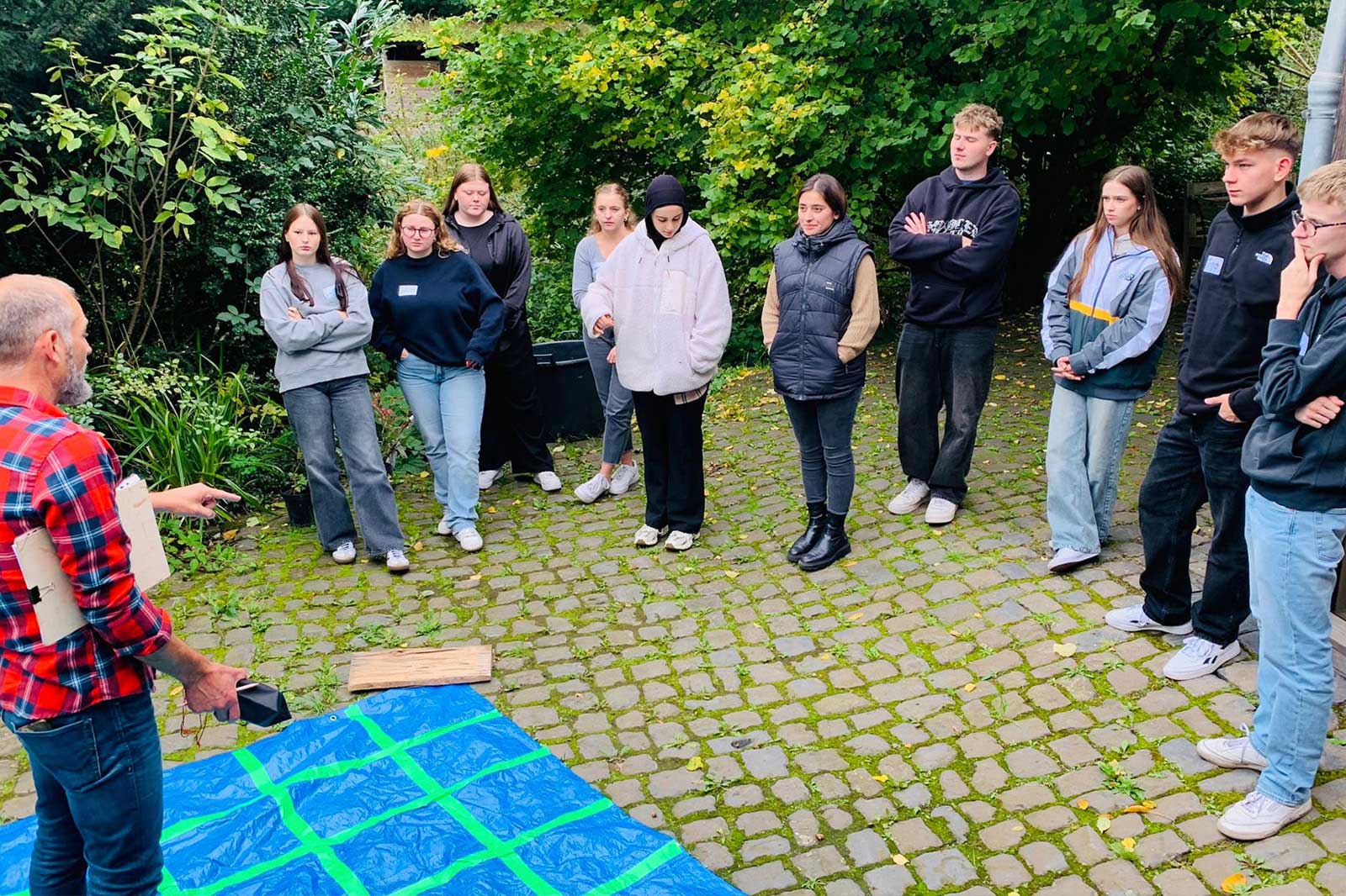 Eine Gruppe von Menschen, die im Freien versammelt ist und aufmerksam einem Sprecher zuhört, in der Nähe einer blauen Plane auf einem Kopfsteinpflaster.