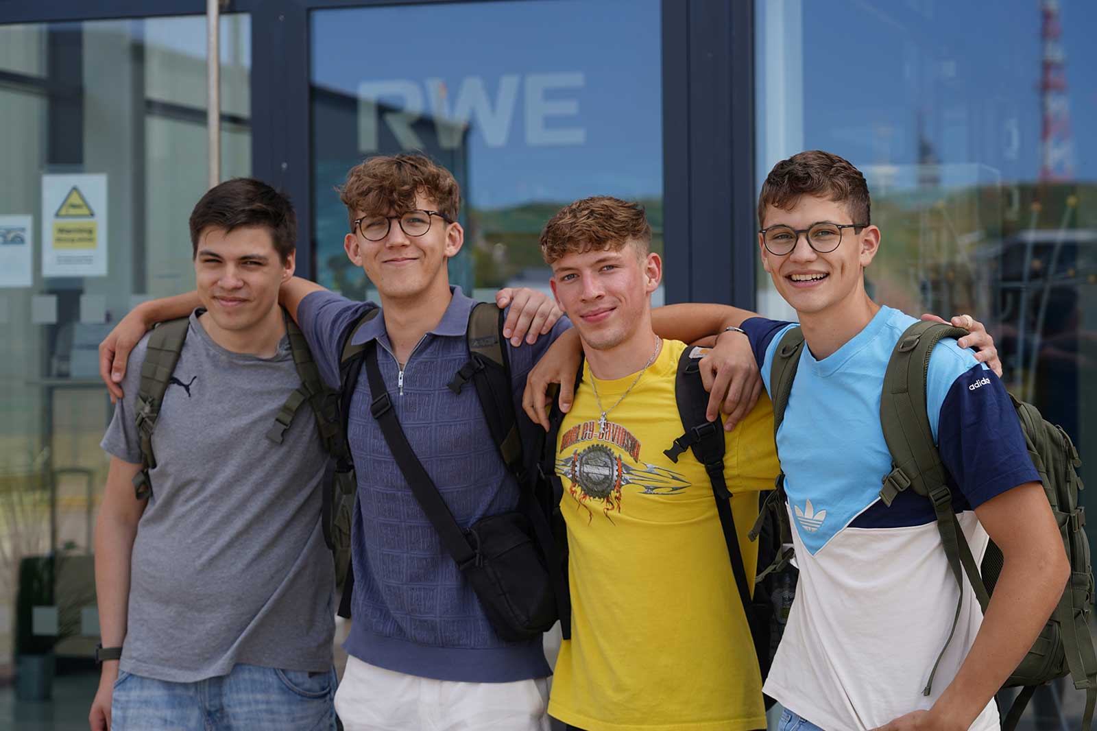 Vier junge Männer posieren zusammen in lässiger Kleidung und mit Rucksäcken vor einem Gebäude mit dem RWE-Logo.