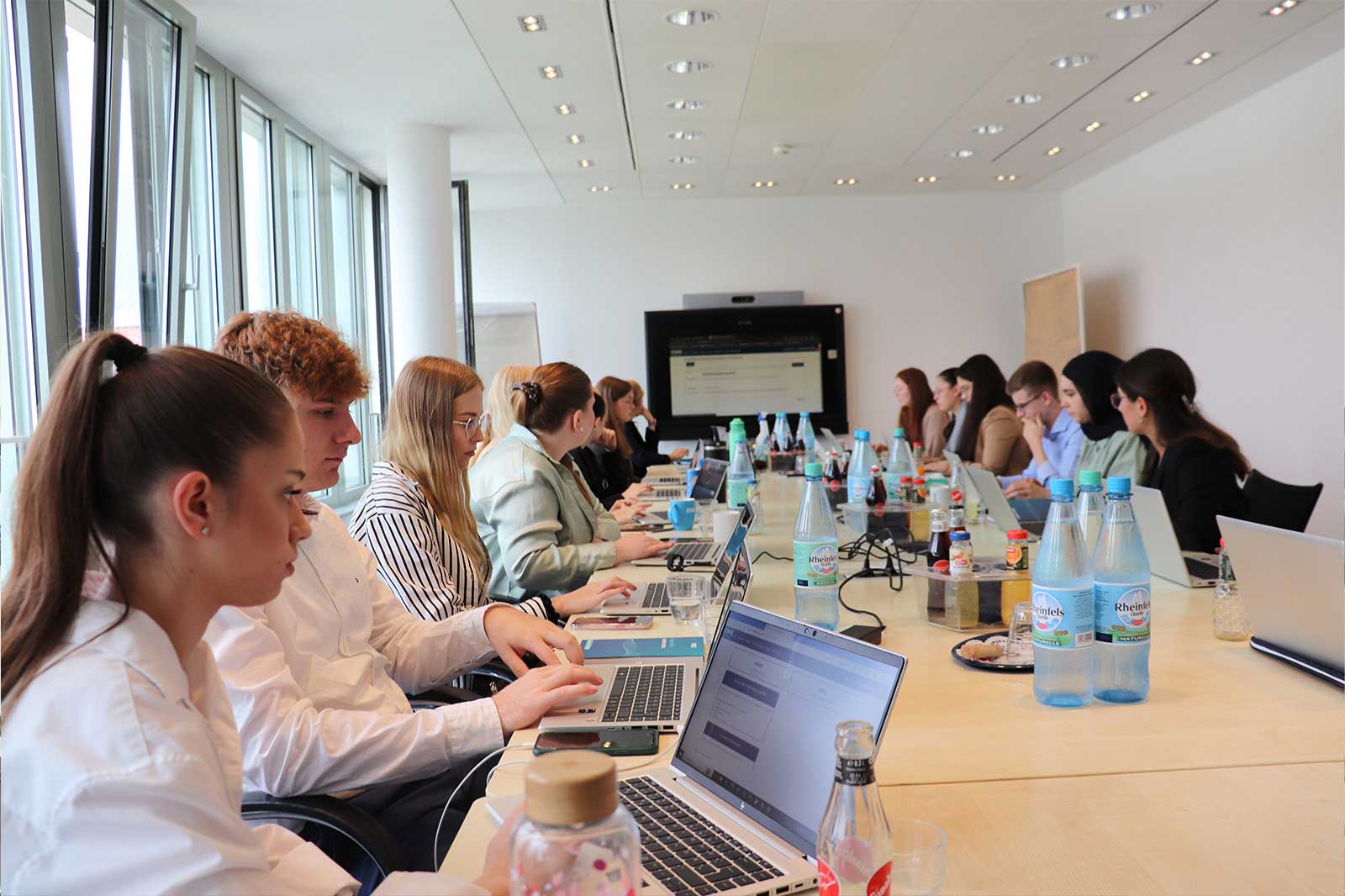 Eine Gruppe von Fachleuten sitzt an einem langen Tisch in einem Besprechungsraum und diskutiert, während Laptops und Erfrischungen um sie herum stehen.
