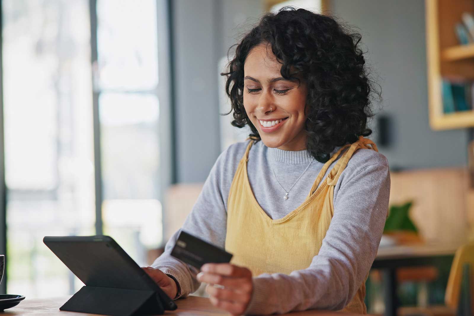 A person with curly hair sits at a table, holding a credit card and using a tablet in a modern café.