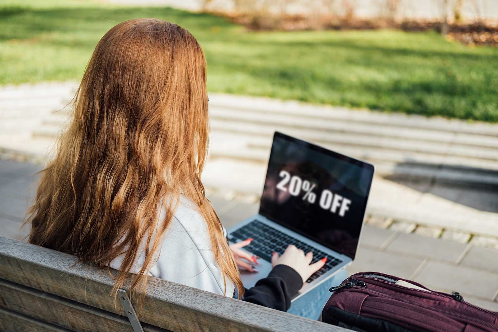 A person with long, wavy, chestnut hair sits on a bench, working on a laptop displaying a 20% off discount.