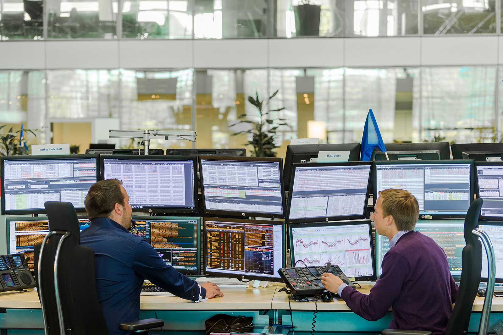Two individuals work at multiple monitors in a modern office featuring plants and a minimalist design.