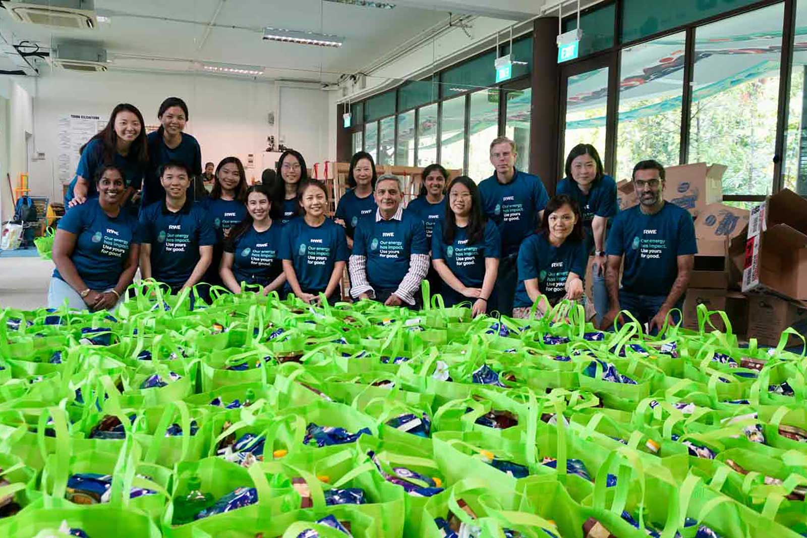 A group of volunteers in blue shirts poses in front of green bags filled with supplies for community support.