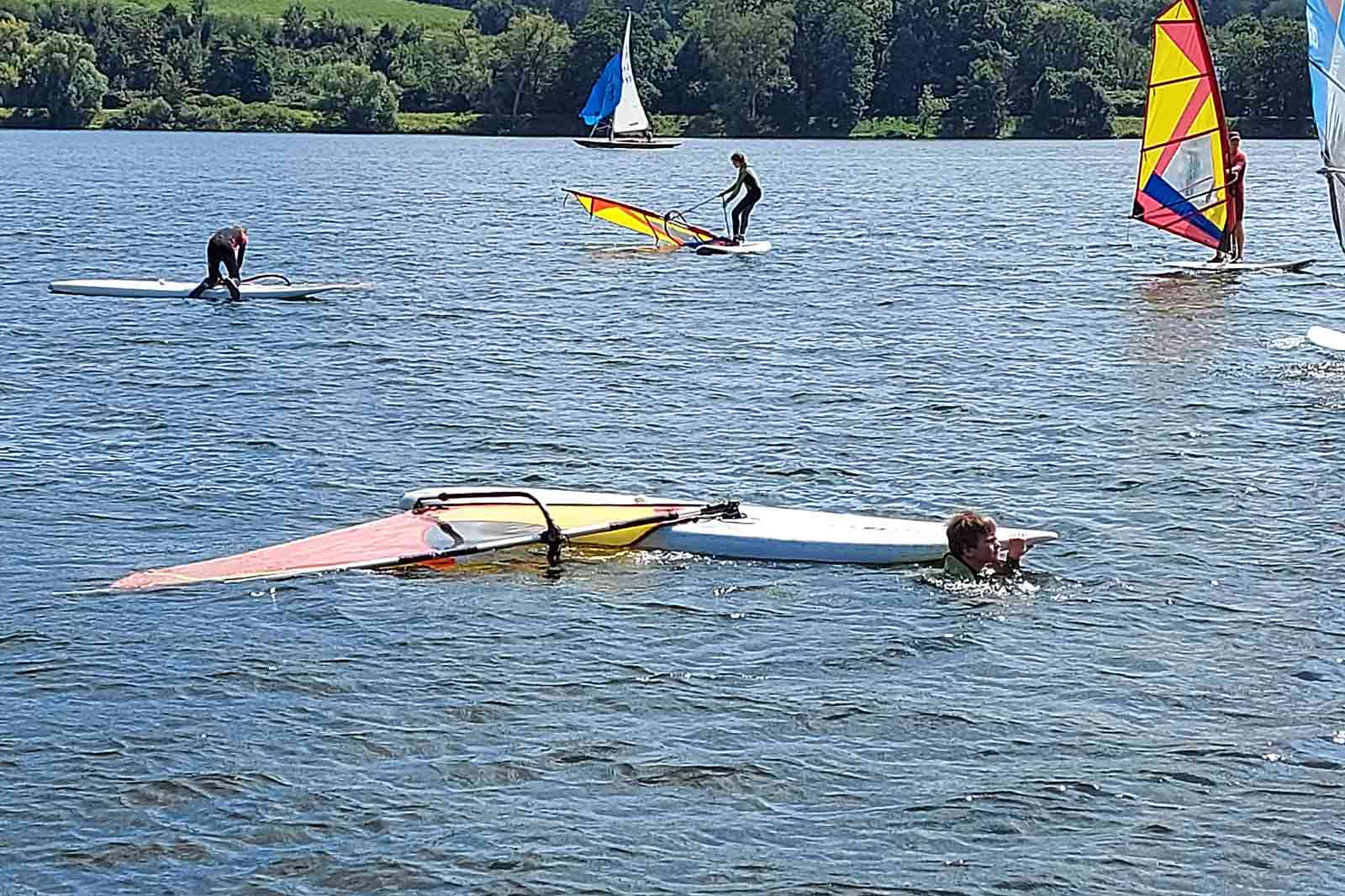 Eine Person im Wasser neben einem umgekippten Windsurfbrett, umgeben von anderen, die auf einem See Windsurfen.
