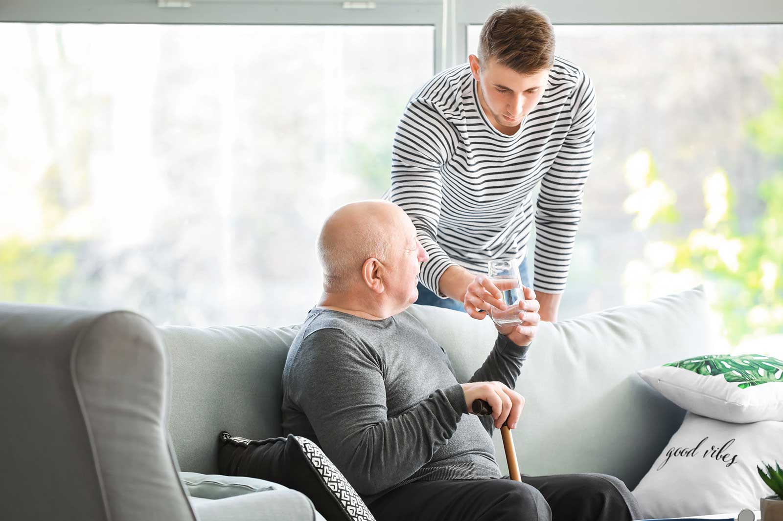 Ein älterer Mann sitzt auf einer Couch und erhält ein Glas Wasser von einem jüngeren Mann in gestreiftem Oberteil.