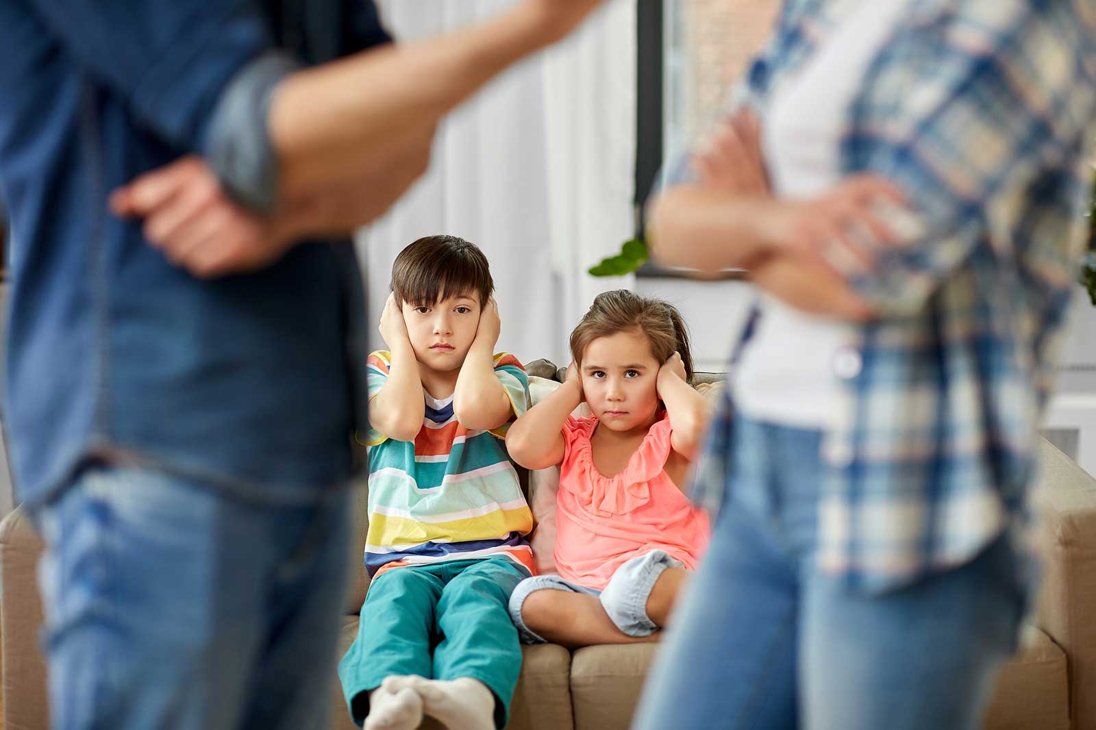 Zwei Kinder sitzen auf einem Sofa und halten sich die Ohren zu, während Erwachsene im Hintergrund streiten.