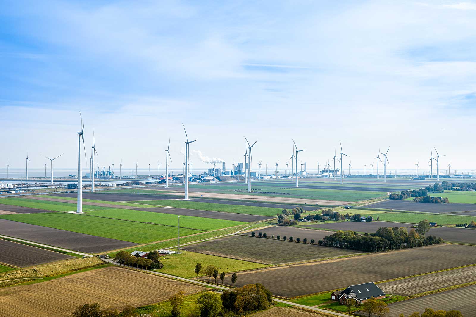 Aerial view of vast fields with wind turbines and a coastal industrial area in the background under a clear blue sky.